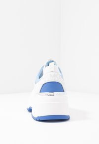 Vita plattformssneakers med blå accenter, med en strukturerad vit häl och dragflik, märkta med "MICHAEL KORS" på sidan.