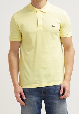 Mand iført en lysegul poloshirt med korte ærmer med et lille grønt logo på venstre bryst og mellemblå jeans.