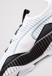 Sneaker bianca con tomaia testurizzata, suola in gomma nera e accenti turchesi. Presenta un design lineare e un tacco pronunciato.