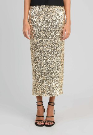 Femme portant une jupe midi en sequins dorés et des sandales noires à talons hauts à brides, debout devant un fond gris uni.