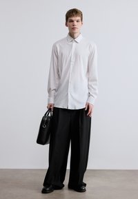 Λευκό πουκάμισο μακρυμάνικο με κουμπωτή πατιλέτα, συνδυασμένο με φαρδιά μαύρη παντελόνια και μαύρη δερμάτινη τσάντα, στέκεται σε ανοιχτόχρωμο δάπεδο.