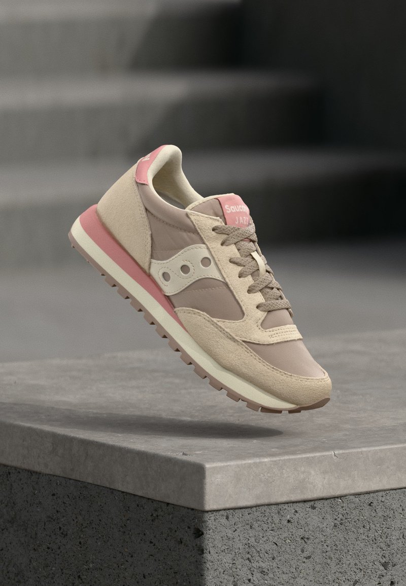 Zapatilla Saucony Jazz en beige y rosa con materiales de ante y tela, flotando sobre un borde de hormigón gris con escalones al fondo.