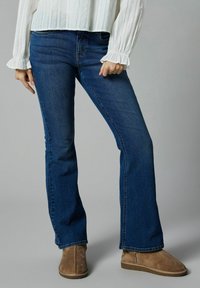 Jean flare en denim bleu avec une texture lisse, comportant des poches avant et une taille mi-haute. Associé avec des bottines en daim beige.
