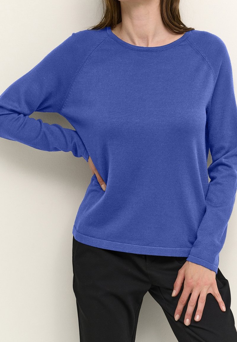 Femme portant un pull bleu à manches longues et col rond, et un pantalon noir, une main sur la hanche et l'autre reposant sur la cuisse.