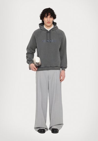 Sudadera gris con un logotipo en el frente, combinada con pantalones de rayas gris claro. El modelo sostiene un pequeño bolso blanco estructurado. Zapatos negros.