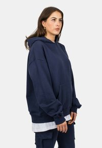 Námořnická modrá oversized mikina, z bavlněné směsi, s přední kapsou, kapucí se stahovací šňůrkou a manžetami s žebrováním, nošena přes bílou vrstvenou košili.