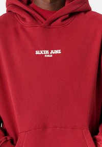 Sudadera roja con un bolsillo frontal y capucha con cordones. Presenta texto blanco que dice "SIXTH JUNE" y "PARÍS". Textura de tela suave.