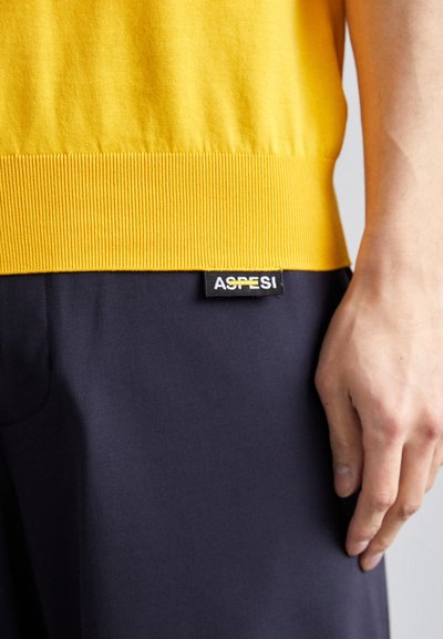 Yellow cotton sweater with ribbed hem, paired with dark navy trousers featuring an ASPI logo tag. Hand resting on trousers, side view.