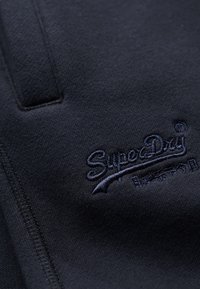 Marineblauwe sweatshirtstof met een soepele textuur, met een donkerblauw borduurlogo waarin "SuperDry" staat, evenals accenten en stikdetails.