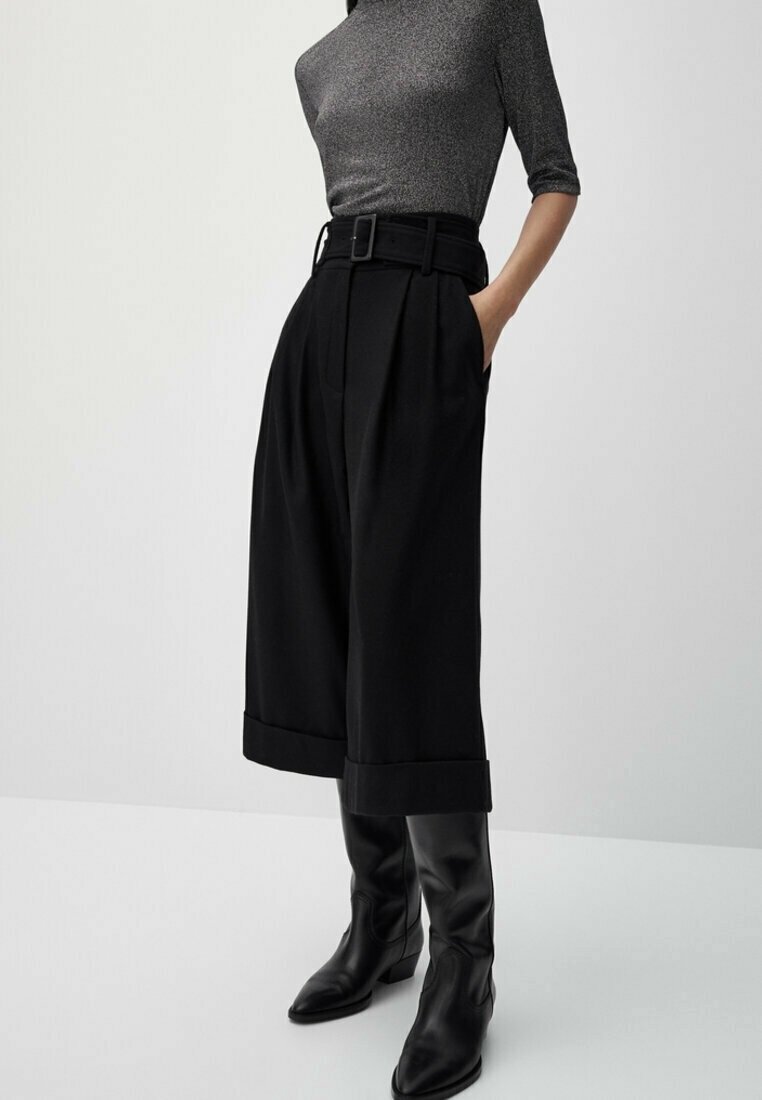 Zwarte high-waisted, wijde broek met plooien, voorzien van een riem en voorzakken, gecombineerd met zwarte leren laarzen tot de knie.