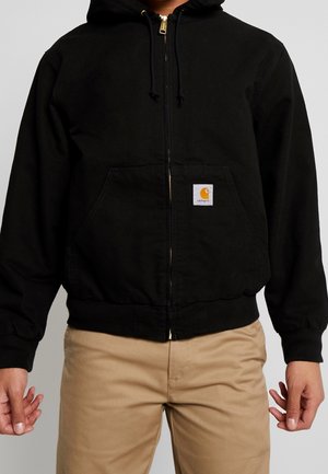 Personne portant un sweat à capuche noir zippé avec poches avant et un petit logo Carhartt, associé à un pantalon beige sur fond gris.