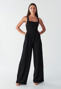 Black jumpsuit with a square neckline, wide-leg silhouette, shoulder straps, and side pockets, made from smooth, lightweight fabric.