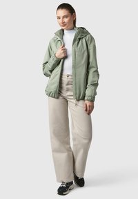 Veste zippée vert clair avec capuche, poignets élastiques et ourlet à cordon de serrage. Portée par-dessus un col roulé blanc et associée à un pantalon beige et des baskets noires.