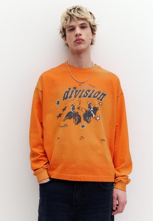 Giovane uomo con capelli biondi che indossa una felpa arancione con cavalieri cowboy e la scritta "The division", in piedi davanti a uno sfondo bianco.