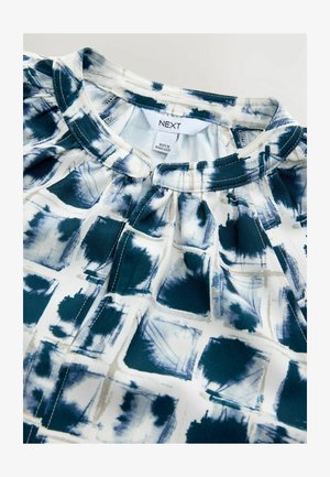 Blouse blanche avec un motif abstrait bleu, dotée d'un col rond, de plis et d'une étiquette au col. Texture de tissu lisse.