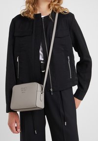 Giacca bomber nera con due tasche frontali con zip, abbinata a una borsa a tracolla grigio chiaro con texture. La giacca presenta un collo rotondo e una vestibilità rilassata.