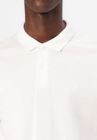Polo bianco realizzato in cotone texturizzato, con un colletto a tre bottoni e un logo ricamato in modo discreto sul lato sinistro del petto.