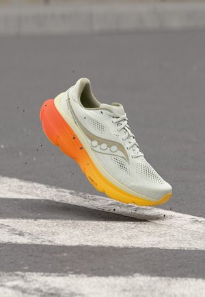 Light gray running shoe with orange-yellow sole hovering above asphalt street with white crosswalk lines.
