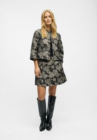 Veste noire à motifs floraux associée à une jupe assortie. Le modèle porte des bottes noires jusqu'aux genoux. Texture lisse, design structuré et manches courtes.