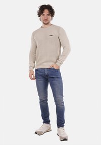 Maglione beige lavorato a maglia con texture a coste, design aderente, scollo rotondo e piccolo logo sul petto, abbinato a jeans di denim blu e sneakers.