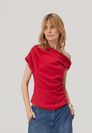 Blondynka w asymetrycznym czerwonym topie i niebieskich dżinsach, patrząca w bok z rękami w kieszeniach na jednolitym jasnym tle.