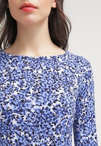 Blå blommönstrad topp med långa ärmar; har en rund halsringning och ett mjukt, texturerat tyg. Designen inkluderar svarta accenter bland de blå blommorna.