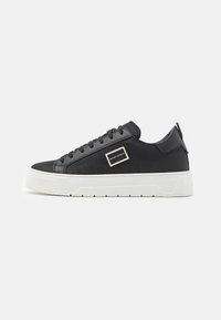 Tenis negri din textile cu un material texturat, sireturi negre plate, talpă din cauciuc albă și un accent de logo metalic pe lateral.