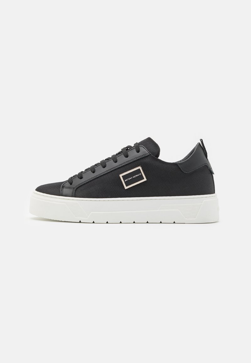 Tenis negri din textile cu un material texturat, sireturi negre plate, talpă din cauciuc albă și un accent de logo metalic pe lateral.