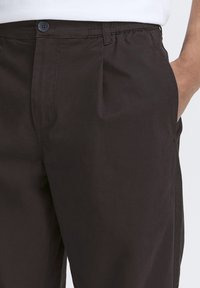 Pantaloni marrone scuro con una texture liscia, dotati di chiusura con bottoni, vita elastica e tasche laterali.