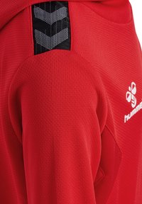 Rød sports-hoodie med tekstureret stof, prydet med et sort og grått chevron-patch på skulderen og hvidt brandlogo på ryggen.