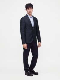 Jeune homme debout portant un costume sombre avec une chemise bleu clair et des chaussures noires sur un fond blanc uni.