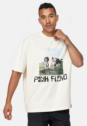 T-shirt à manches courtes couleur crème avec un graphique de vache et le texte "PINK FLOYD" en noir gras sous l'image. Tissu en coton doux.