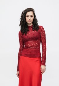 Dolga rdeča čipkasta majica z dolgimi rokavi in oprijetim krojem, z visokim ovratnikom in valovitim zaključkom spodaj, v kombinaciji z gladko rdečo satenasto krilo.
