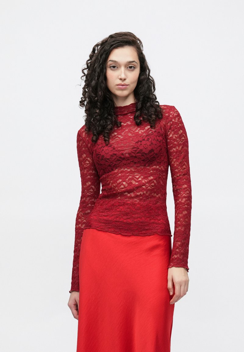 Dolga rdeča čipkasta majica z dolgimi rokavi in oprijetim krojem, z visokim ovratnikom in valovitim zaključkom spodaj, v kombinaciji z gladko rdečo satenasto krilo.