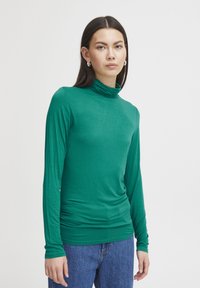 Grüner langärmeliger Rollkragenpullover aus glattem Stoff, mit einer taillierten Silhouette und Raffungen an der Taille, kombiniert mit blauen Jeans.