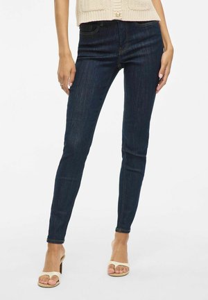 Dark blue skinny jeans made of denim, featuring a high waist, five pockets, and a smooth texture. Worn with beige heeled sandals.