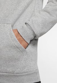 Sudadera gris con un bolsillo tipo canguro, puños acanalados y una tela suave y texturizada. Primer plano mostrando una mano parcialmente metida en el bolsillo.