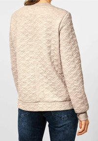 Prošívaná béžová mikina s žebrovanými manžetami a výstřihem, zdobená geometrickým vzorem a s postranními rozparky na lemu pro větší pohodlí.