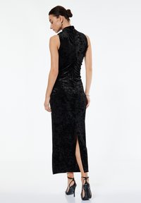 Svart sammetsklänning med hög krage, ärmlös design, rynkdetaljer och en sidöppning. I kombination med svarta rem-högklackade skor.