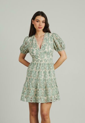 Mujer de pie luciendo un vestido de encaje verde pálido, de manga corta y escote en V, con un patrón floral y mangas abullonadas, contra un fondo claro y liso.