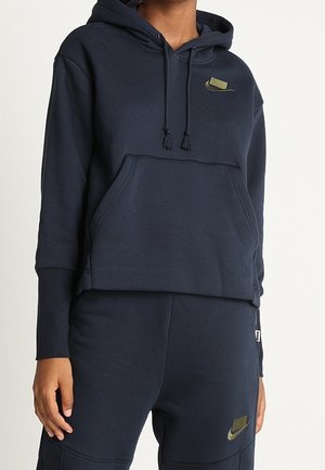 Osoba ubrana w granatową bluzę z kapturem i sznurkami oraz pasujące spodnie, oba z małymi oliwkowozielonymi logo Nike swoosh.