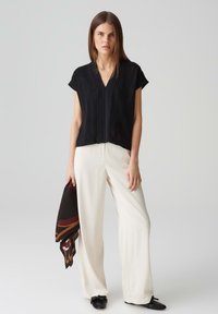 Top nero a maniche corte con scollo a V e lavorazione a maglia, abbinato a pantaloni larghi color crema. La modella tiene una sciarpa scura a fantasia.
