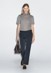 Grijze kortärmige coltrui, donkerblauwe op maat gemaakte broek, bruine instappers en een kleine bruine handtas, gepresenteerd tegen een neutrale achtergrond.