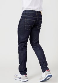 Pantalones vaqueros de denim azul oscuro con un corte slim, costuras amarillas, bolsillos en la parte trasera y combinados con zapatillas deportivas grises y blancas.