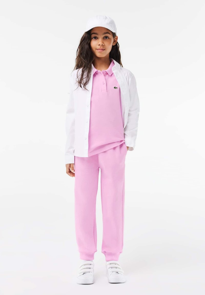 Polo rose, chemise blanche à boutons, et joggings roses. Casquette blanche et baskets. Des textures lisses et des couleurs contrastées créent un look superposé.