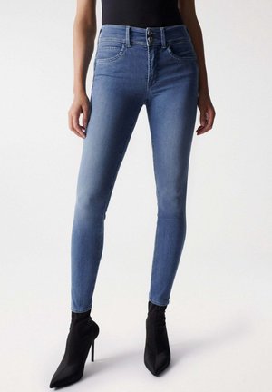 Jeans skinny blu in denim, con vita alta, due tasche frontali e chiusura con bottone, abbinati a stivaletti neri con punta affilata.