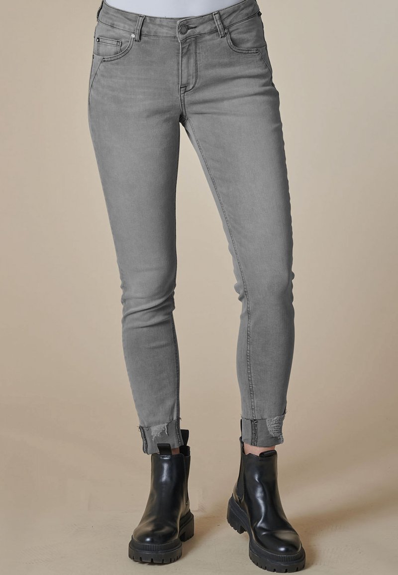 Frau trägt hellgraue Slim-Fit-Jeans mit ausgefransten Säumen und schwarze klobige Stiefeletten, steht auf beigem Boden.