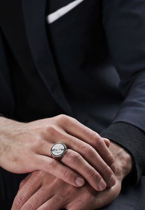 Silvrig kompassdesignring med en polerad yta, rund band och intrikata detaljer på toppen, bärs på en hand med mörk kostym.