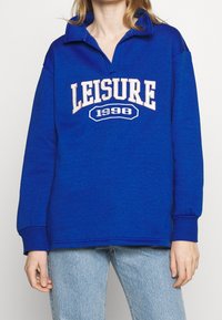 Blå halsringad sweatshirt med krage, med texten "LEISURE 1998" i krämfärgad färg över framsidan. Mjuk tyg och ribbade manschetter.