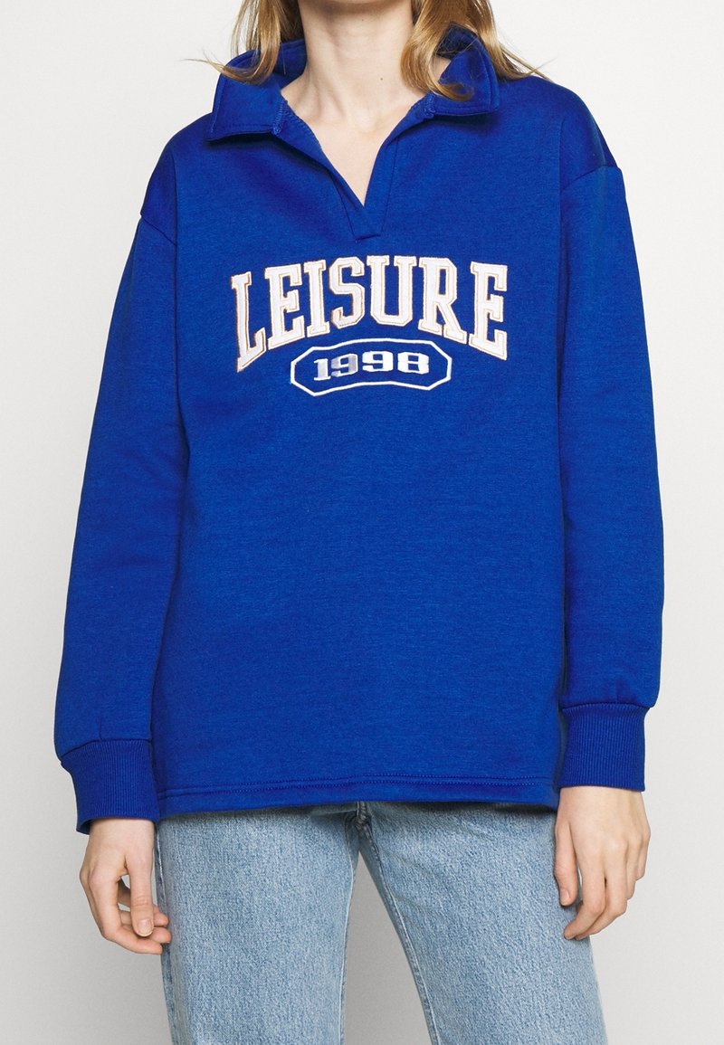 Blå halsringad sweatshirt med krage, med texten "LEISURE 1998" i krämfärgad färg över framsidan. Mjuk tyg och ribbade manschetter.